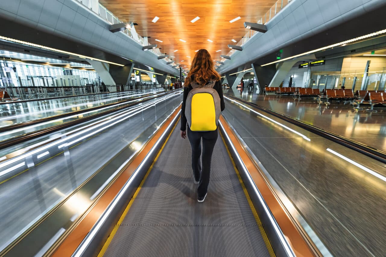 One woman traveller with backpack go on moving walkway travolator in new Hamad International Airport in Doha, Qatar. Back view